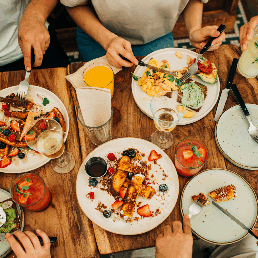 Overhead view of a wooden table with people eating brunch, including French toast with berries, scrambled eggs with avocado, and waffles. Glasses of juice, wine, and cocktails are also on the table.
